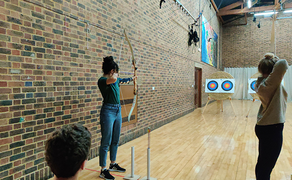 Woman in sports hall holding a bow and aiming at a target in the distance